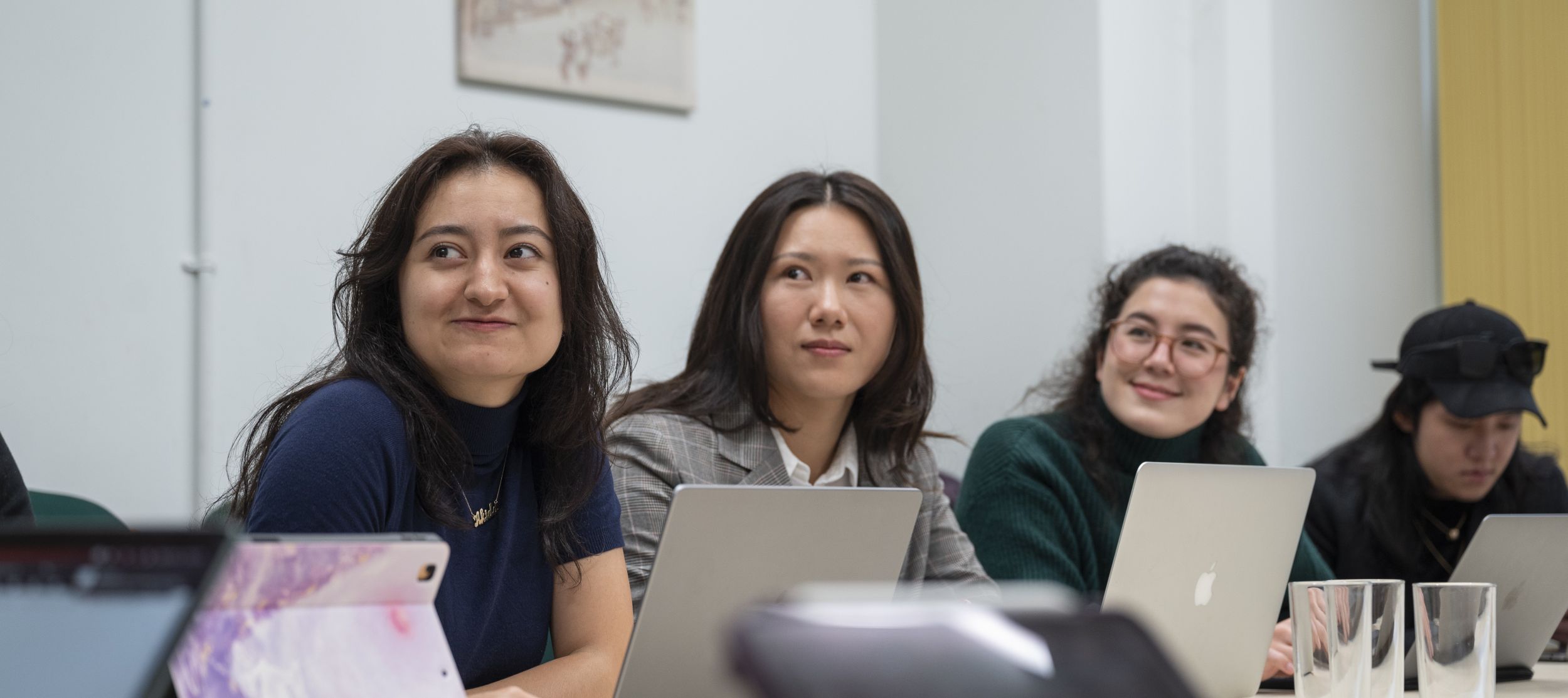 Four students sitting at a table in a seminar room with laptops and tablets in front of them