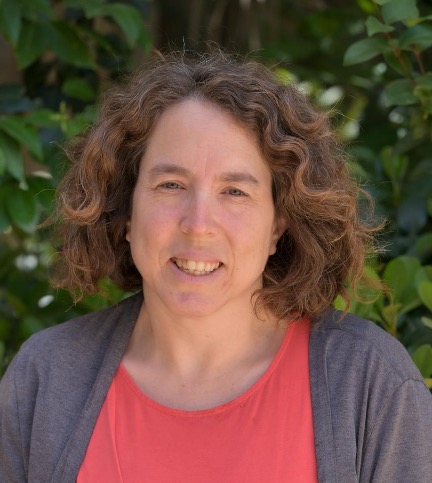 Tali has curly brown shoulder-length hair and she is smiling at the camera. She's wearing a salmon pink t shirt under a grey cardigan.  
