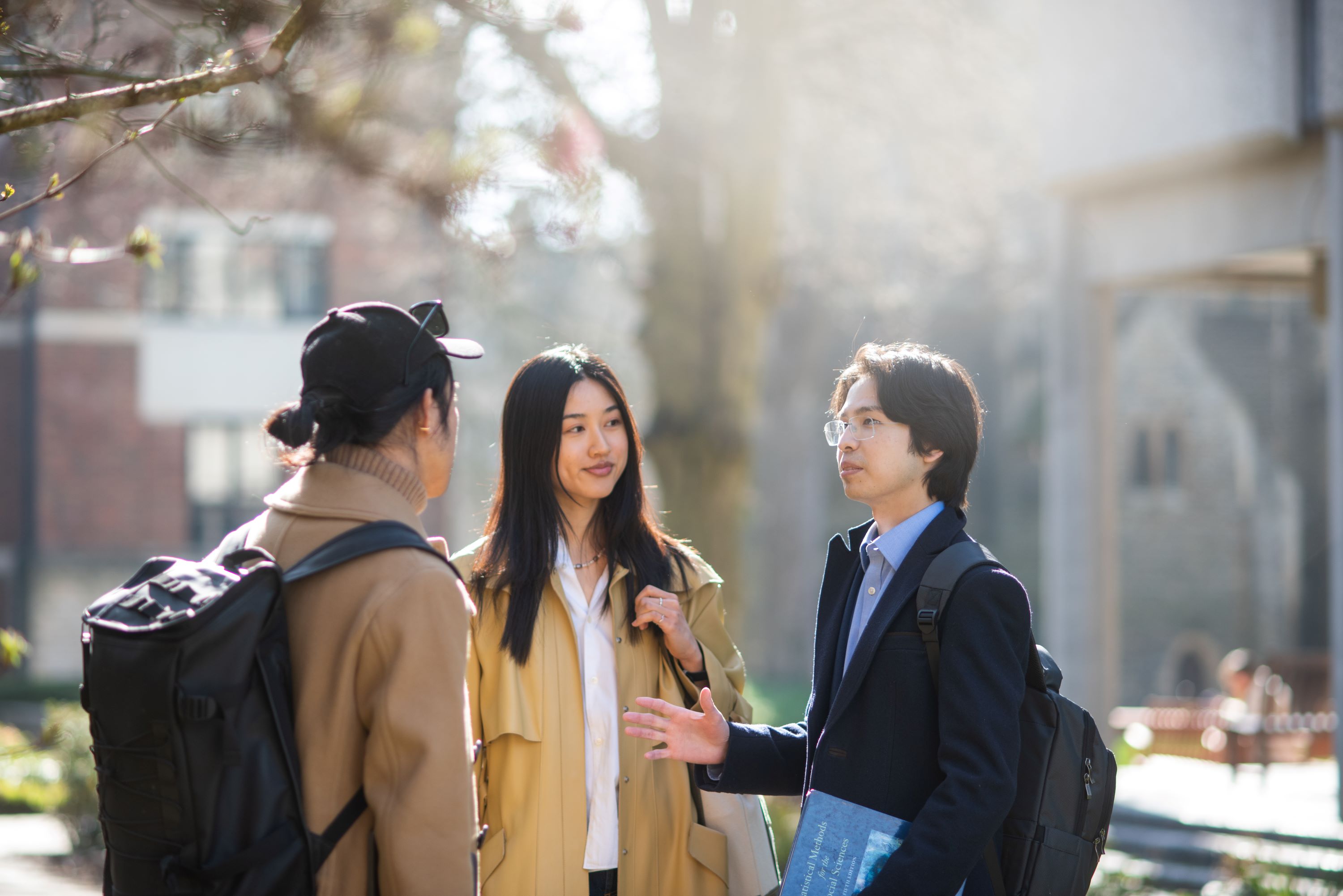 Three students standing outside under a large tree. They are wearing light winter coats and carrying backpacks.