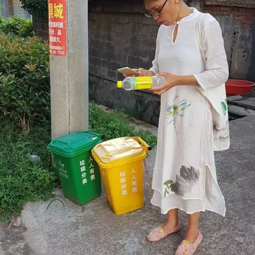 A lady wearing a long white dress holding a plastic bottle in her hand as she stands next to one yellow, and one green recycling bin. 