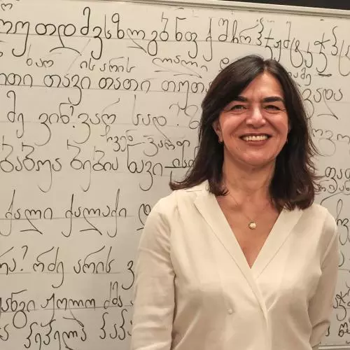 Lia Chokoshvili has shoulder-length dark hair. She is wearing a white v-neck blouse and standing in front of a whiteboard covered in Georgian script.