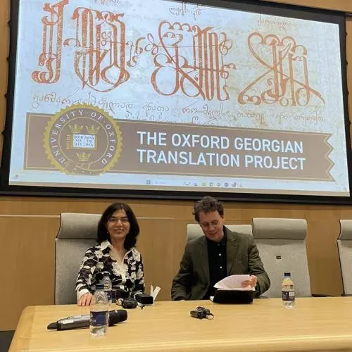 Lia Chokoshvili and Oliver Matthews sitting at a large wooden desk. Lia is smling at the camera and Oliver is looking down at a book he has opened, smiling.