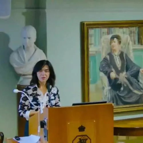 Lia Chokoshvili is standing and speaking behind a brown lectern. She is wearing a black and white patterned blazer