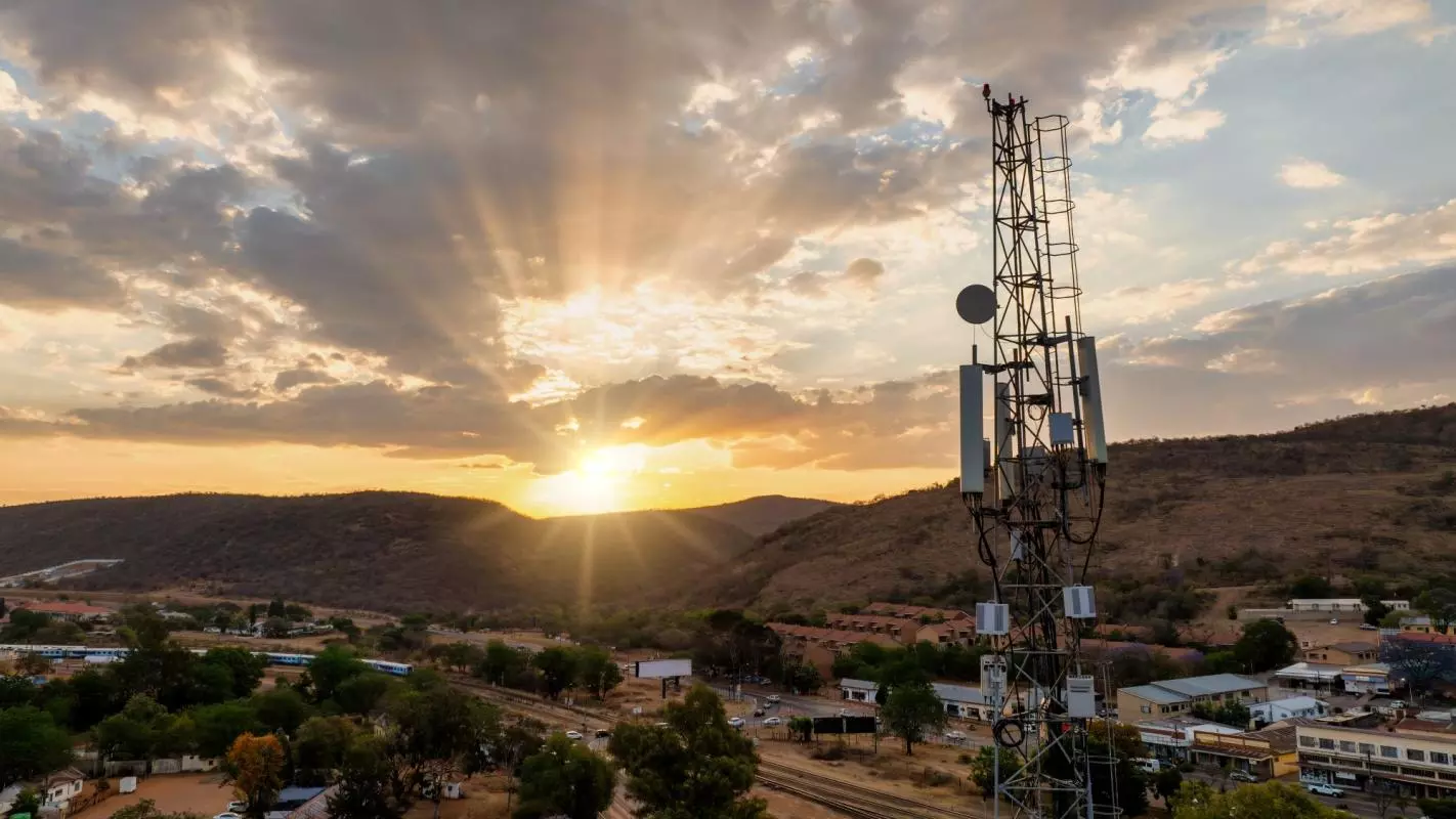 An aerial view of a cellular antenna at sunset, in front of a town, railway line and some hills.