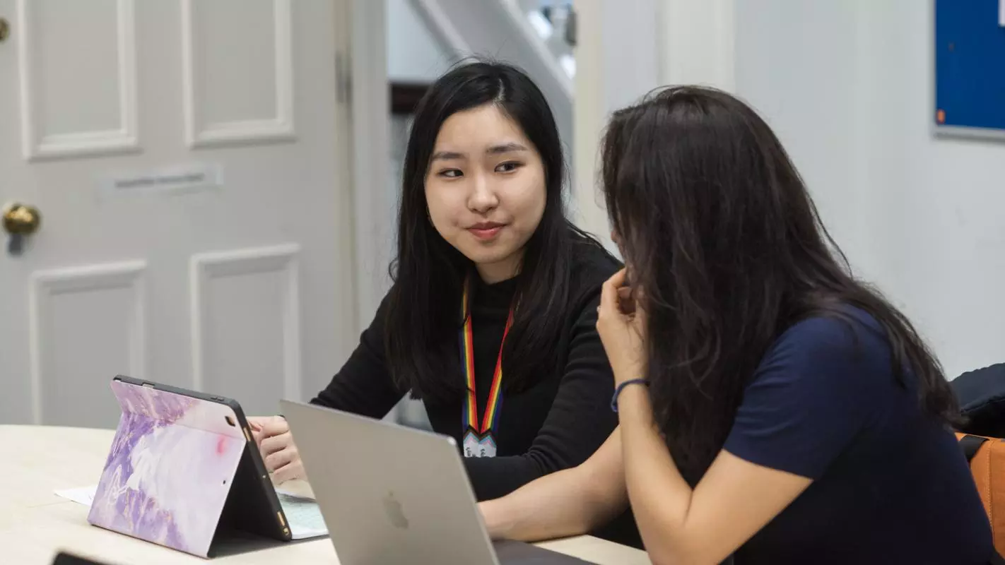 Two students sitting at a desk with laptops, chatting to each other. 