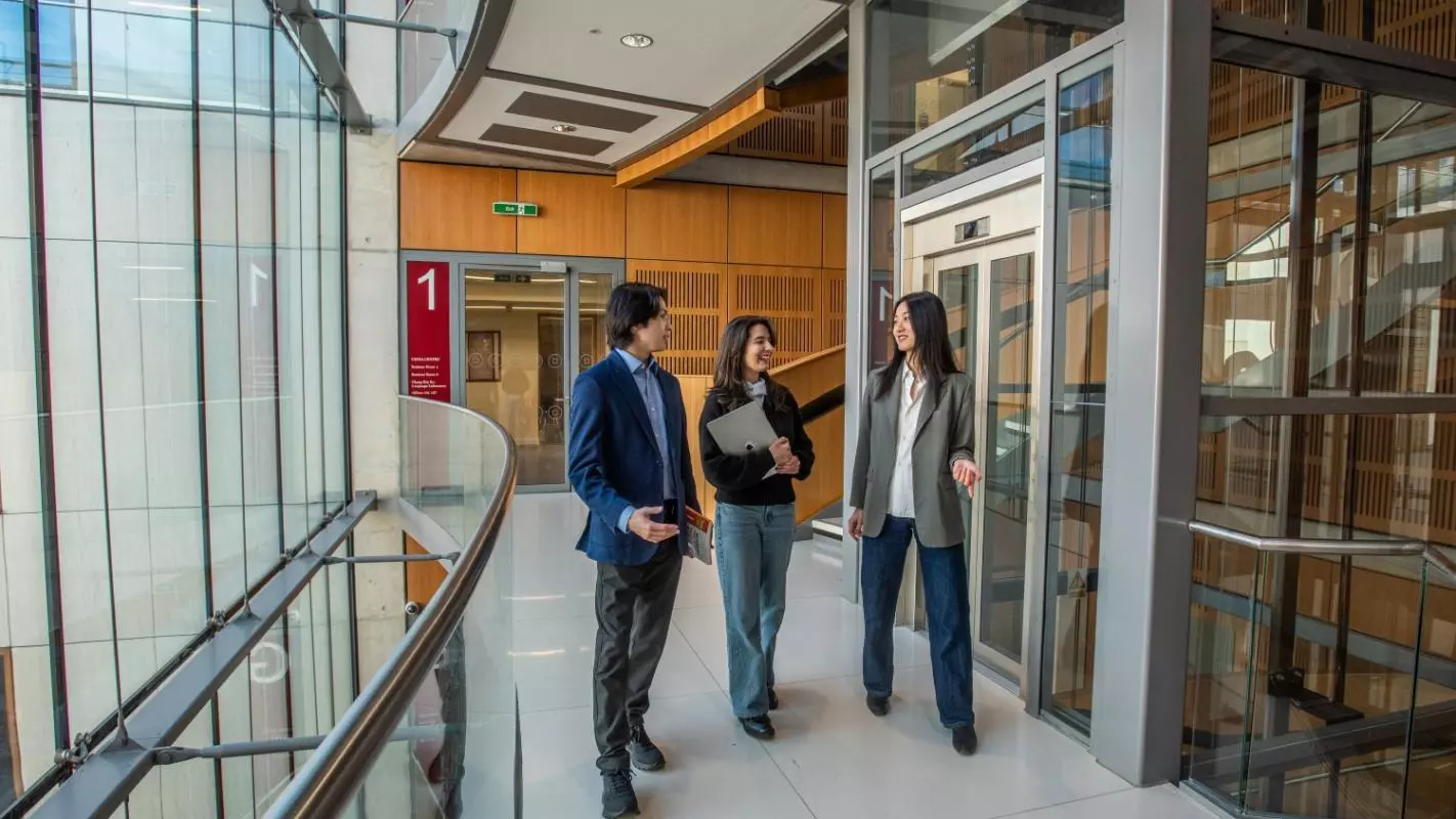 Three students walking through a corridor of a modern department building, surrounded by large glass windows. 