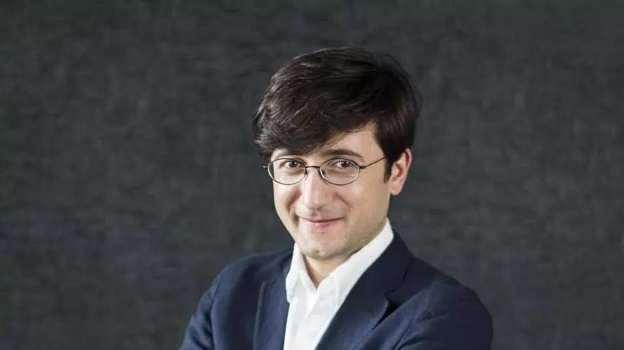 Dr Maziashvili has short dark hair. He is wearing a navy blue suit with a white shirt, standing with his arms folded and smiling