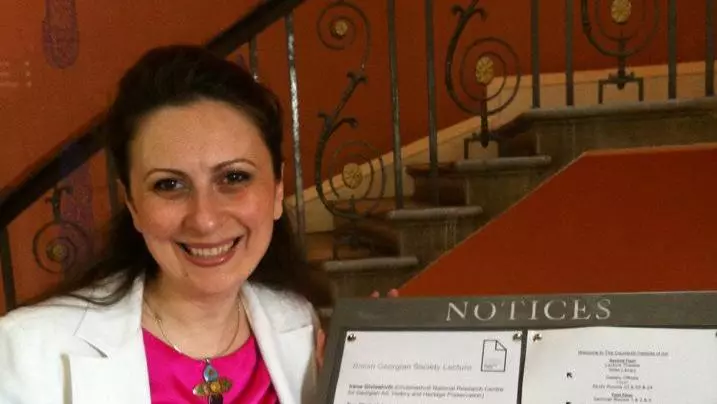 Dr Giviashvili is wearing a bright pink top and white blazer. She is standing next to a board of notices