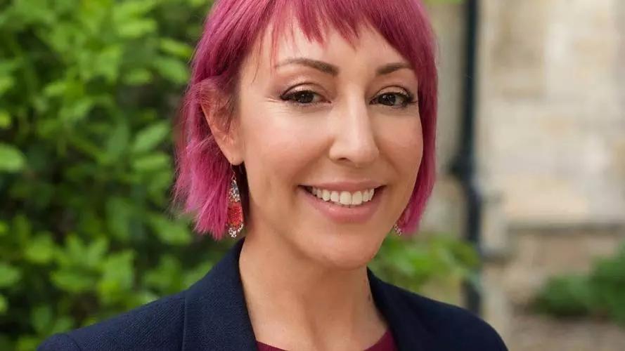 Janey has pink hair in a short bob with fringe. She's wearing a wine red top with navy blue jacket