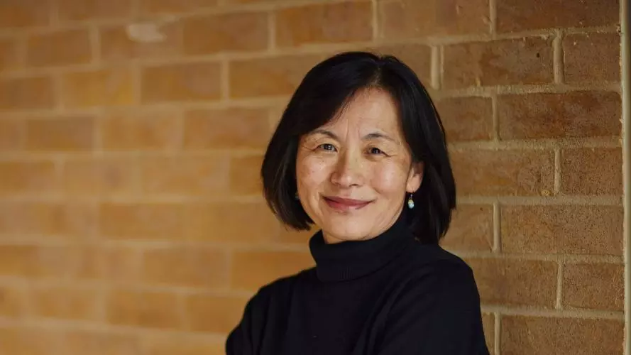 Professor Sako has dark bobbed hair and is wearing a black top. She is leaning against and wall with her arms folded, smiling