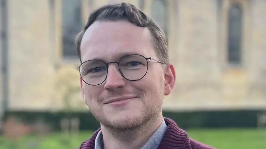 Daniel has short brown hair, a beard and is wearing glasses. He's wearing a blue shirt under a maroon zip up sweater. He's standing in front of an Oxford college. 
