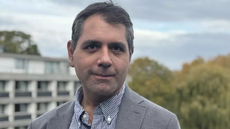 Professor Naseemullah has short dark hair and is wearing a blue and white patterned shirt with a grey blazer. He is standing on a rooftop in front of modern concrete buildings and some trees