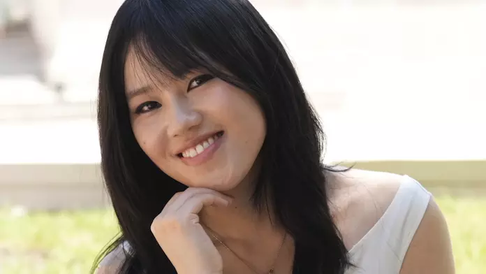  Mingdan has long black hair, is resting her chin on her hand, and is smiling at the camera. She's wearing a sleeveless white top