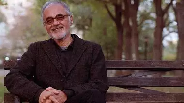 Mahmood Mamdani, Herbert Lehman Professor of Government and African Studies at Columbia University. He is sitting on a wooden bench surrounded by trees. 