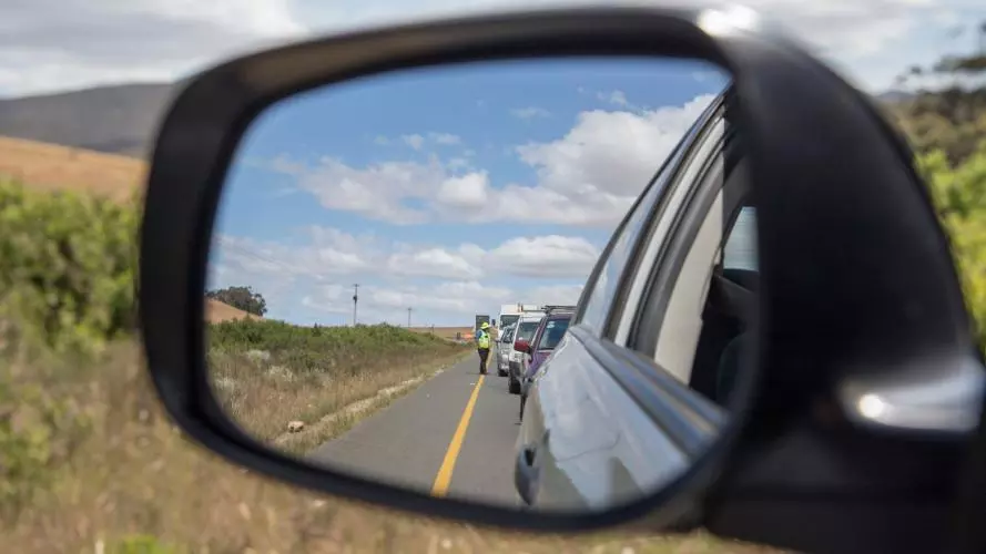 A close-up of a car wing mirror, which is reflecting a queue of traffic being monitored by someone in a high-vis jacket.
