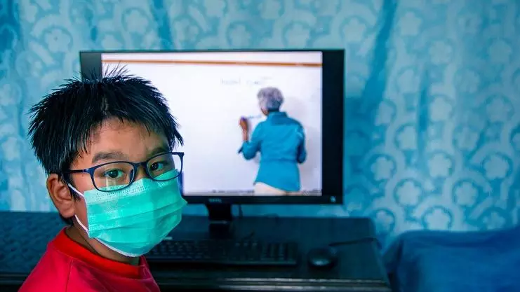 A boy with black hair and glasses wearing a aquamarine face mask. He is watching an education programme on TV.