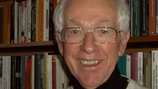 Alan has grey hair, glasses and is smiling at the camera. He's wearing a black shirt under a cream coloured jumper. He's standing in front of a bookcase.