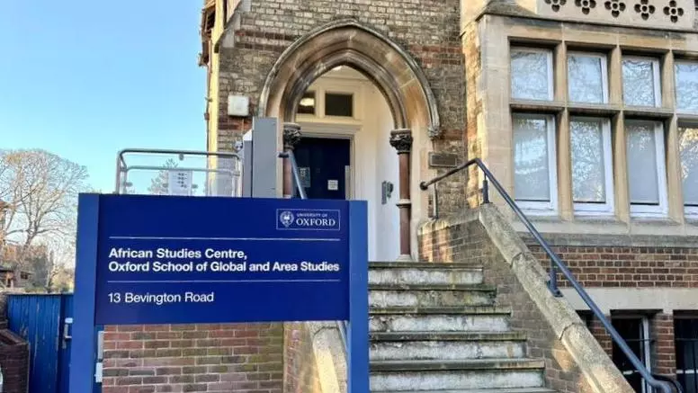 The African Studies Centre is a tall building made of light brick. It has large bay windows and a staircase leading to an arch before a porch. There is a new, dark blue sign outside the front with the Centre name. 