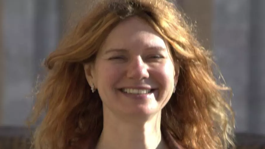 Anna has long auburn hair and is smiling at the camera. she's wearing a beige knitted top with a brown jacket. 