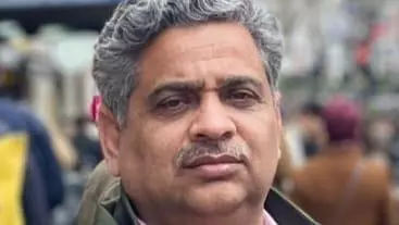 Professor Raja has grey curly hair and a moustache. He is wearing a pink shirt under a grey jumper, and a dark green coat.  