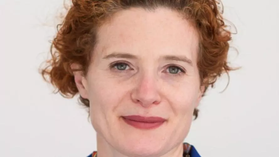 Anna Lora-Wainwright has curly auburn hair that she has tied back. She is wearing a royal blue shirt with a necklace made from various multicoloured beads. 