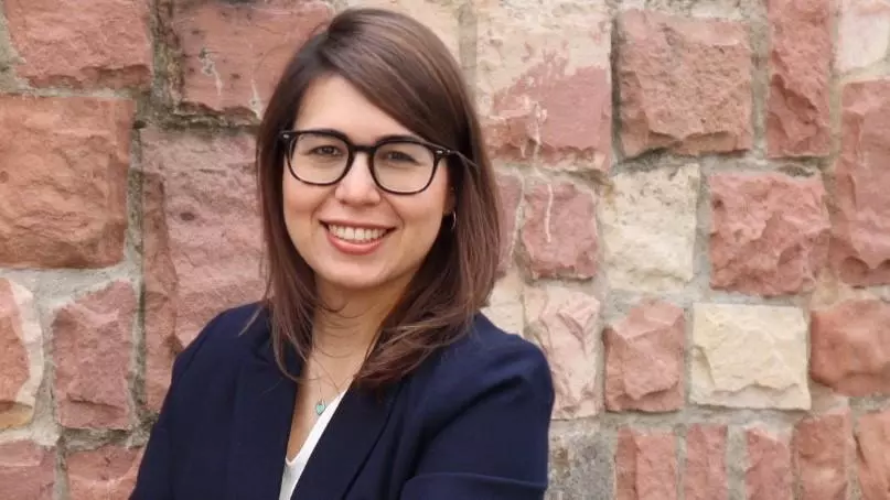 Maryhen had shoulder length brown hair, is wearing glasses and smiling at the camera. She's wearing a white top under a navy blue jacket. She's standing against a stone wall. 