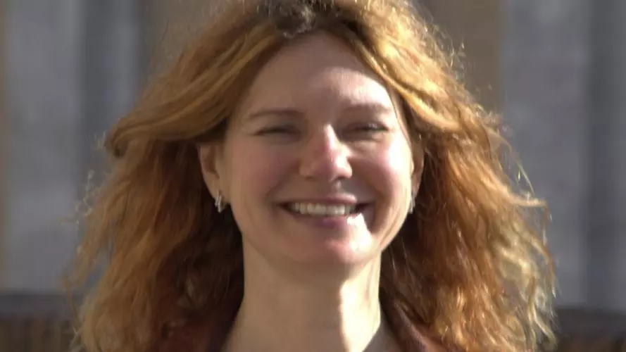 Anna has long auburn hair and is smiling at the camera. she's wearing a beige knitted top with a brown jacket.