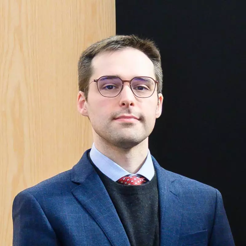 Matthew has short brown hair and is wearing glasses. He's dressed in a smart pale blue shirt with red tie, dark navy jumper and a royal blue blazer.
