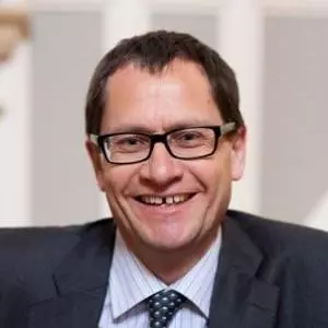 Roger Goodma has short dark hair and dark glasses. He is wearing a shirt, tie and dark blazer