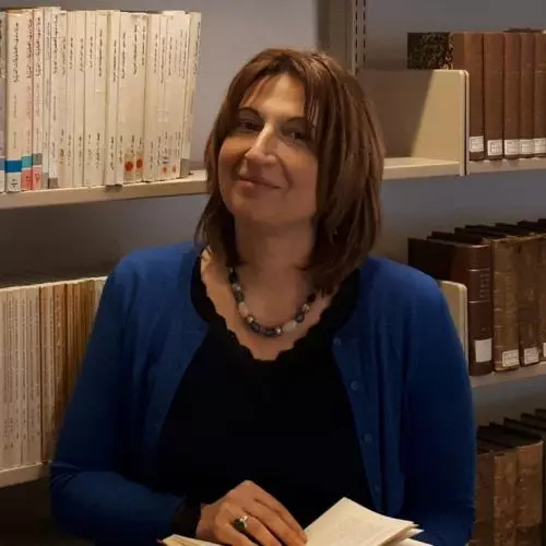 Tamara Pataridze has shoulder-length brown hair and is wearing a dark dress with a blue cardigan. She is holding a book open and standing in front of bookshelves
