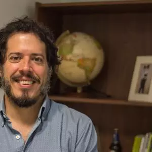 Gabriel has curly black hair and a black beard. He's wearing a pale blue shirt and is standing in front of a shelf with a globe and framed picture.