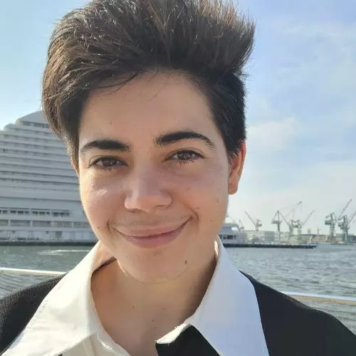 Pan has short dark hair. They are wearing a black and white shirt with a large collar, standing in front of a harbour