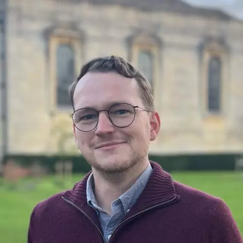 Daniel has short brown hair, a beard and is wearing glasses. He's wearing a blue shirt under a maroon zip up sweater. He's standing in front of an Oxford college. 