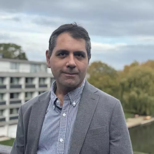 Professor Naseemullah has short dark hair and is wearing a blue and white patterned shirt with a grey blazer. He is standing on a rooftop in front of modern concrete buildings and some trees