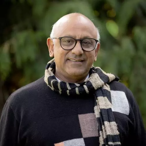 Premesh has grey hair, dark rimmed glasses and is wearing a blue and grey scarf over a navy blue sweater.  There is a blurred green background resembling trees behind him. 