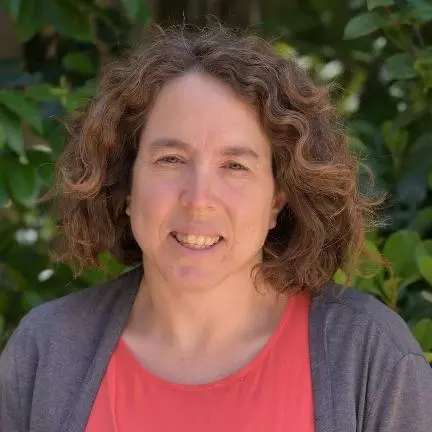 Tali has curly brown shoulder-length hair and she is smiling at the camera. She's wearing a salmon pink t shirt under a grey cardigan.  