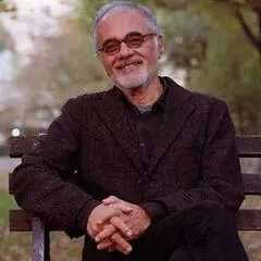 Mahmood Mamdani, Herbert Lehman Professor of Government and African Studies at Columbia University. He is sitting on a wooden bench surrounded by trees. 