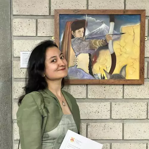 akida anwar has long dark hair and wearing a pale green dress with cropped jacket. She is holding a certificate and standing next to her framed painting. The painting shows an archer on horseback and aiming their bow and arrow. There is a smoking volcano behind the archer on horseback.