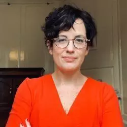 Maria has short dark hair and glasses, and is wearing a bright orange v-neck top