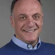Othon has short grey hair and smiling broadly at the camera. He's wearing a sky blue shirt under a dark blue jumper 