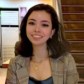 Eliza has shoulder length brown hair and brown eyes. She is wearing a dark blue silk shirt, brown checked blazer and gold jewellery. She is smiling at the camera. 