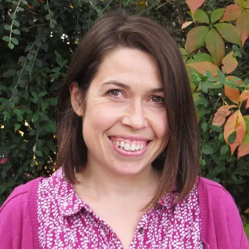 Jo Pready has shoulder length brown hair. She is wearing a pink patterned blouse and pink cardigan and smiling. 