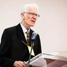 Arthur Stockwin has grey hair and wearing glasses. He's standing at a podium speaking into a microphone. He's wearing a white shirt, tie and smart black jacket. 