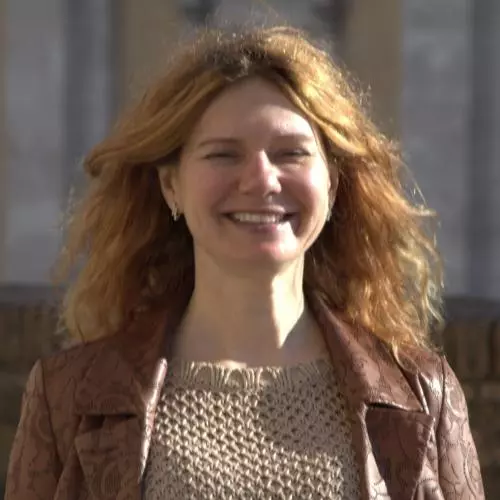 Anna has long auburn hair and is smiling at the camera. she's wearing a beige knitted top with a brown jacket.