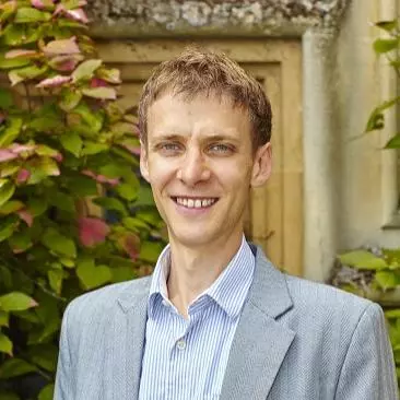 Justin has short light brown hair and is smiling at the camera. He's wearing a shirt, with vertical blue and white stripes, under a grey jacket. 