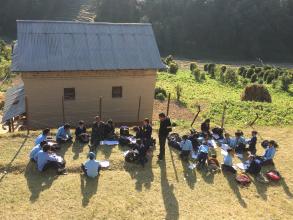 School children sitting on grass outside a school building in a rural area