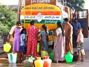 Group of people collecting water from a tank, some are carrying brightly coloured pots with additional pots on the ground near their feet