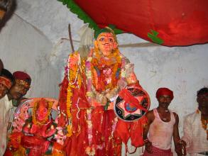 Statue wearing red robes and holding a shield with four people in the background wearing white shirts, some are wearing red hats 