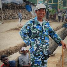 A man wearing a blue, green and white patterned jacket and trousers and beige cap. He's standing up holding a wooden stick, about 1metre long. 