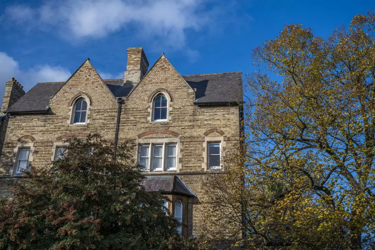 Two joined brown-brick buildings against a blue sky, surrounded by trees and hedges
